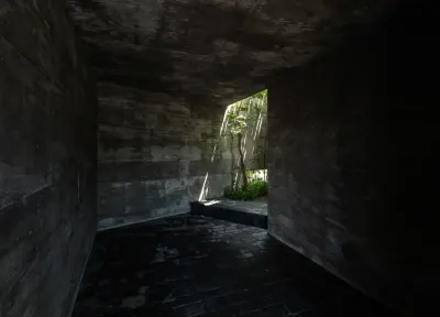 Pasillo de hormigón en penumbra que lleva hacia un patio con un árbol y luz de sol al fondo.