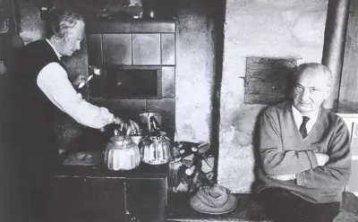 Martin Heidegger y Elfride Heidegger en la cocina de su cabaña: ella atiende las pavas junto a la estufa de azulejos; él, sentado, mira.