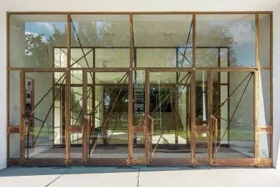 Acceso vidriado de un edificio de Alvar Aalto: puertas con marcos de metal cobrizo y tiradores en diagonal, con una escalera al fondo.