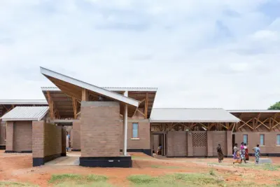 Maternity Waiting Village: casas bajas de ladrillo con techos de chapa y estructura de madera a la vista; unas mujeres caminan por el patio de tierra.