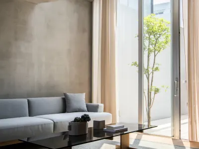 Living de un departamento en Tokio: sillón gris, mesa baja de vidrio y roble, paredes de hormigón y una ventana alta con cortinas livianas que da a un patio con un árbol.