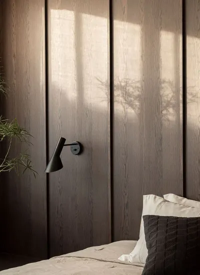 Sol de la tarde sobre una pared revestida en madera oscura, con la sombra de una planta, junto a una cama con almohadones claros y una lámpara de pared negra.