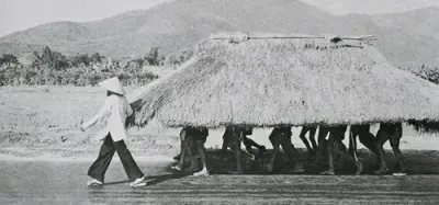 Foto en blanco y negro de un grupo de personas que caminan cargando un techo de paja; bajo su sombra solo se les ven las piernas.