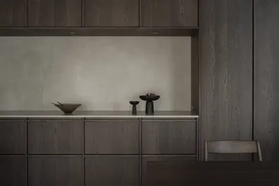 Mueble bajo de madera oscura con un nicho iluminado sobre revoque claro, donde hay un cuenco y dos piezas de cerámica, y una silla de madera al costado.