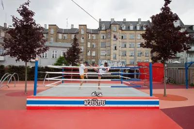 Un ring de boxeo tailandés en la plaza roja de Superkilen, con dos personas entrenando, árboles y edificios de vivienda detrás.