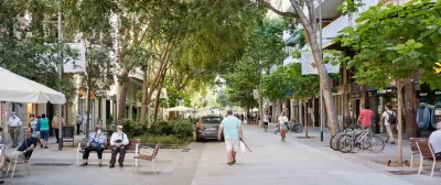 Calle peatonal arbolada en Sant Antoni, Barcelona: gente sentada en bancos, una terraza de café, bicicletas y personas caminando.