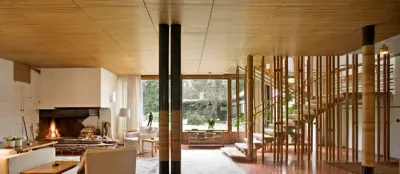 Living de la Villa Mairea: techo de listones de madera, columnas oscuras, una chimenea blanca encendida a la izquierda y una escalera rodeada de varas de madera a la derecha.